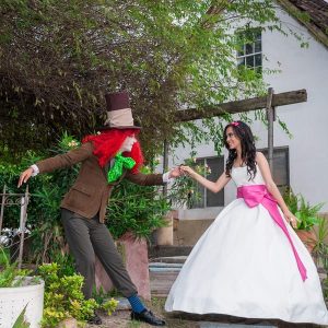 Hermosa Quinceañera recreando una escena de Alicia en el país de las Maravillas junto con el señor del Sombrero en un bello jardín con una cabaña al fondo, fotografía realizada por Click Fotografía en Río Bravo